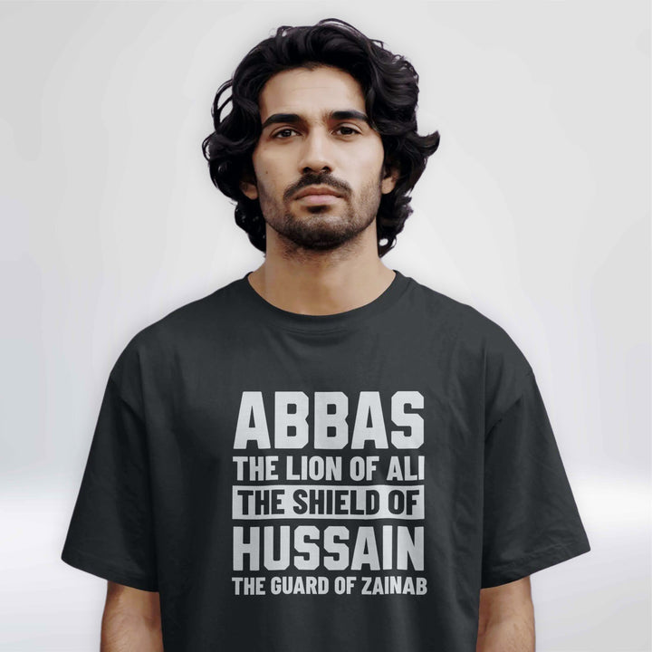 Black T-shirt with bold white text reading "ABBAS – The Lion of Ali, The Shield of Hussain, The Guard of Zainab" hanging on a minimal studio hanger.

