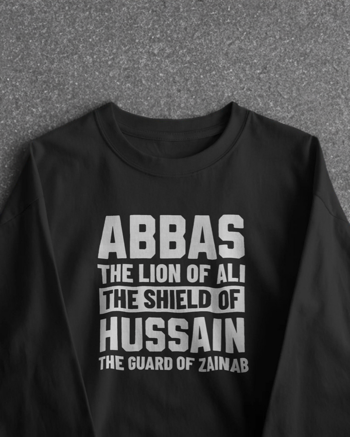 Black Full Sleeves T-shirt with bold white text reading "ABBAS – The Lion of Ali, The Shield of Hussain, The Guard of Zainab" hanging on a minimal studio hanger. 
