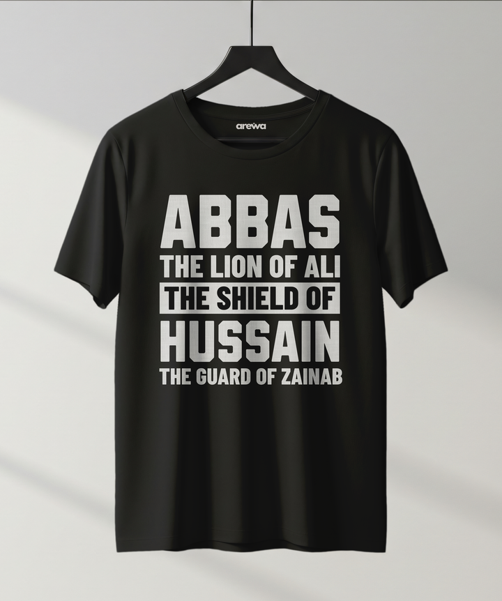 Black T-shirt with bold white text reading "ABBAS – The Lion of Ali, The Shield of Hussain, The Guard of Zainab" hanging on a minimal studio hanger.

